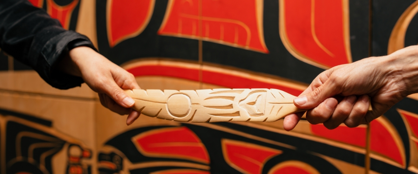 hands exchanging an indigenous carved wooden artwork in front of a tapestry with black and red indigenous artwork on it