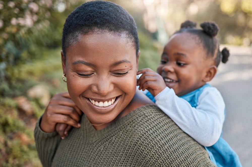 Smiling Black woman outdoors with her daughter on her back and holding on to her shoulder, also smiling