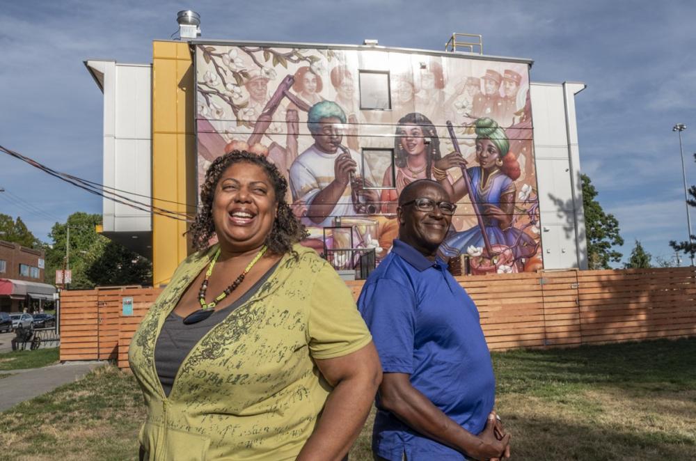 Hogans Alley image of two people standing outdoors smiling in front of a painted mural in Hogans Alley Vancouver BC