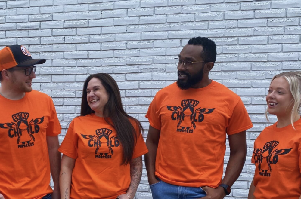 two men and two women wearing orange t-shirts for Orange Shirt Day, smiling at each other, with a white brick wall in the background