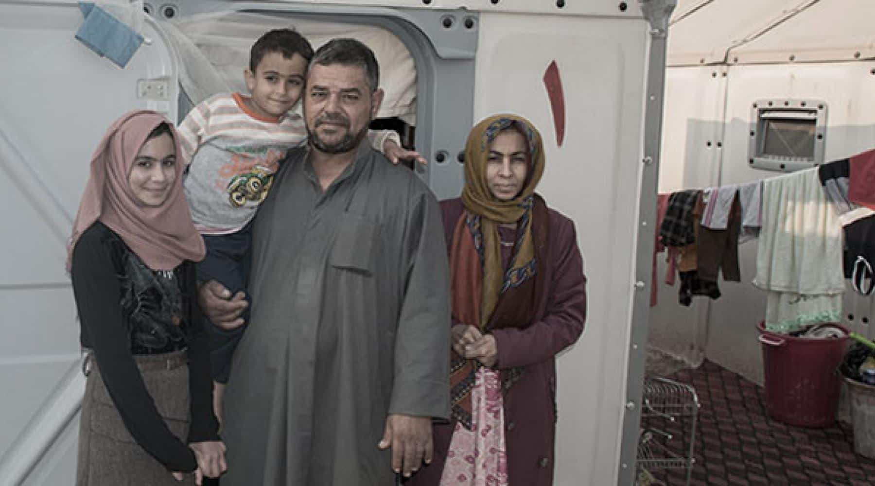 Family standing outside a temporary shelter with laundry hanging nearby.