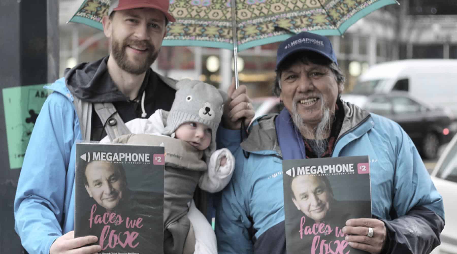 Two people holding Megaphone magazines while standing under an umbrella on a rainy city street, with a baby in a carrier.