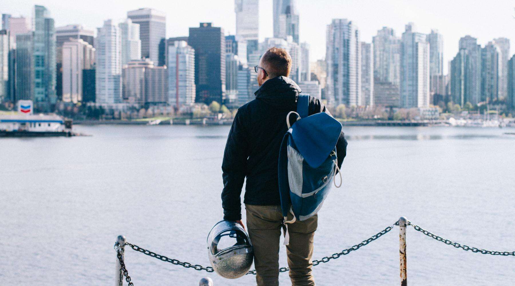 Person with a backpack holding a helmet while looking out at the Vancouver skyline across the water.