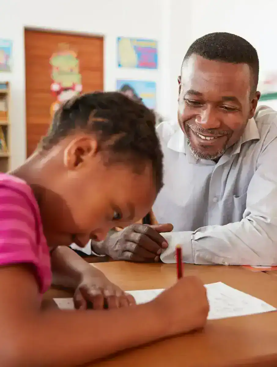 Teacher smiling while watching a young student write on paper at a desk in a classroom