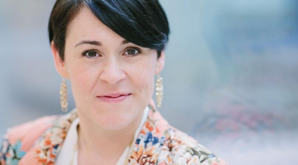 Professional headshot of a woman with short dark hair wearing earrings and a patterned blazer.