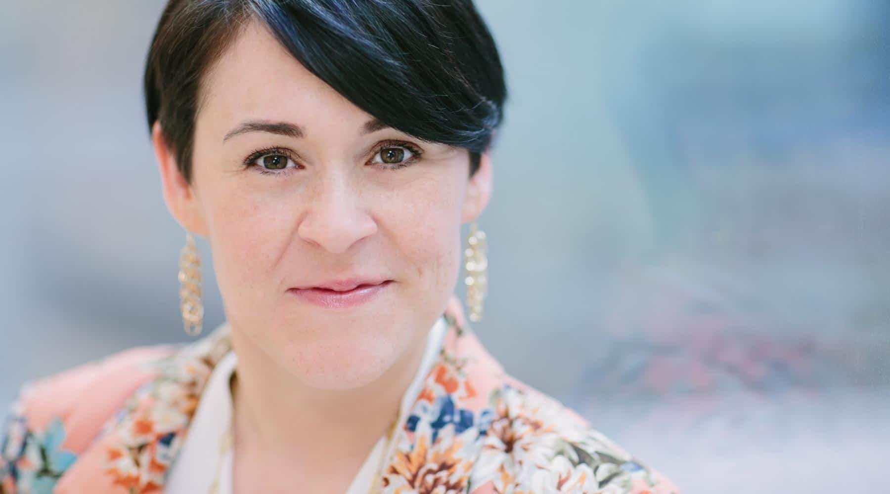 Professional headshot of a woman with short dark hair wearing earrings and a patterned blazer.