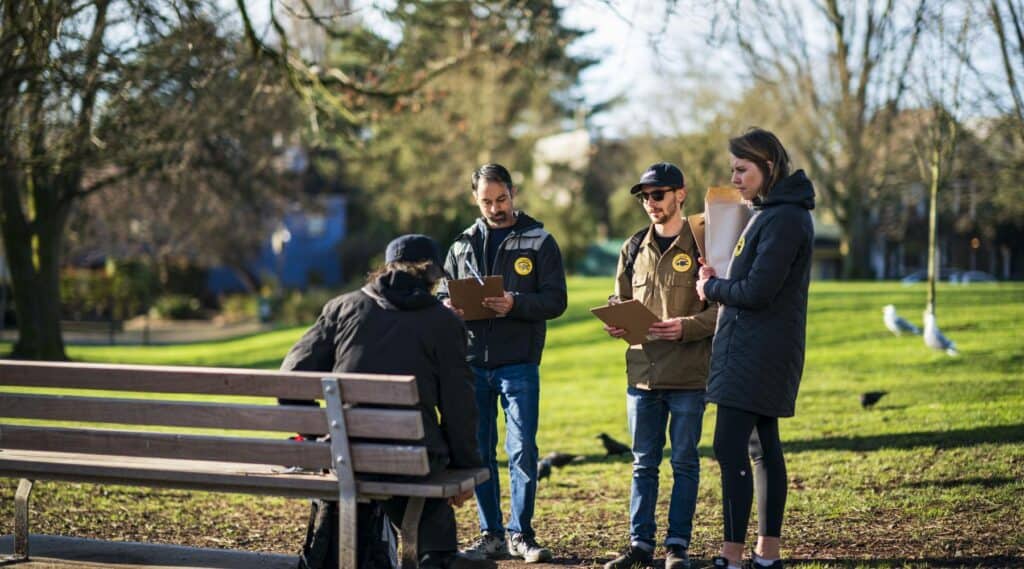 Outreach workers speaking with a person sitting on a park bench while taking notes on clipboards.