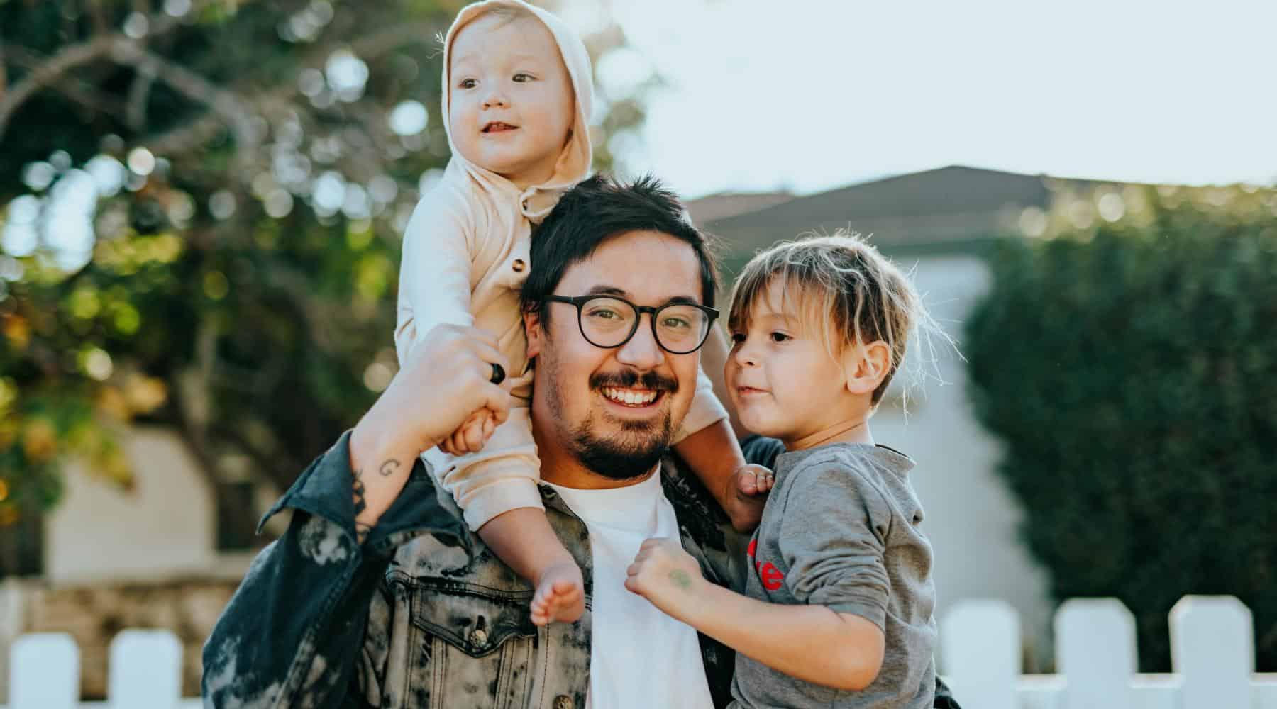 Father holding two young children outdoors in front of a home