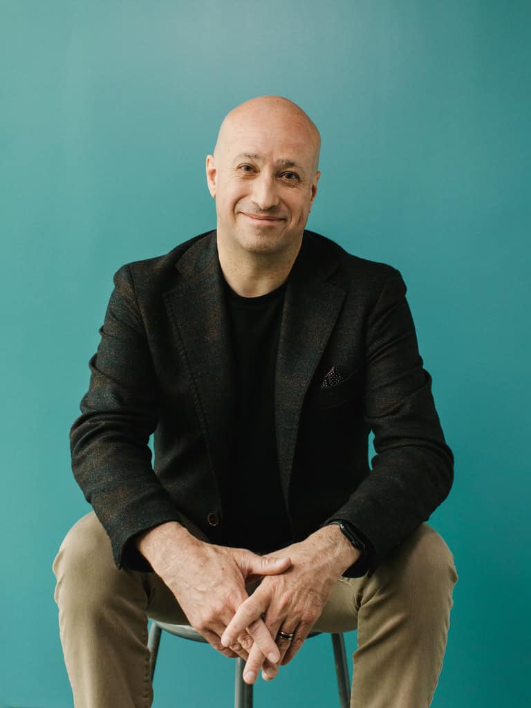 Smiling bald man wearing a dark blazer and black shirt, seated on a stool against a teal background