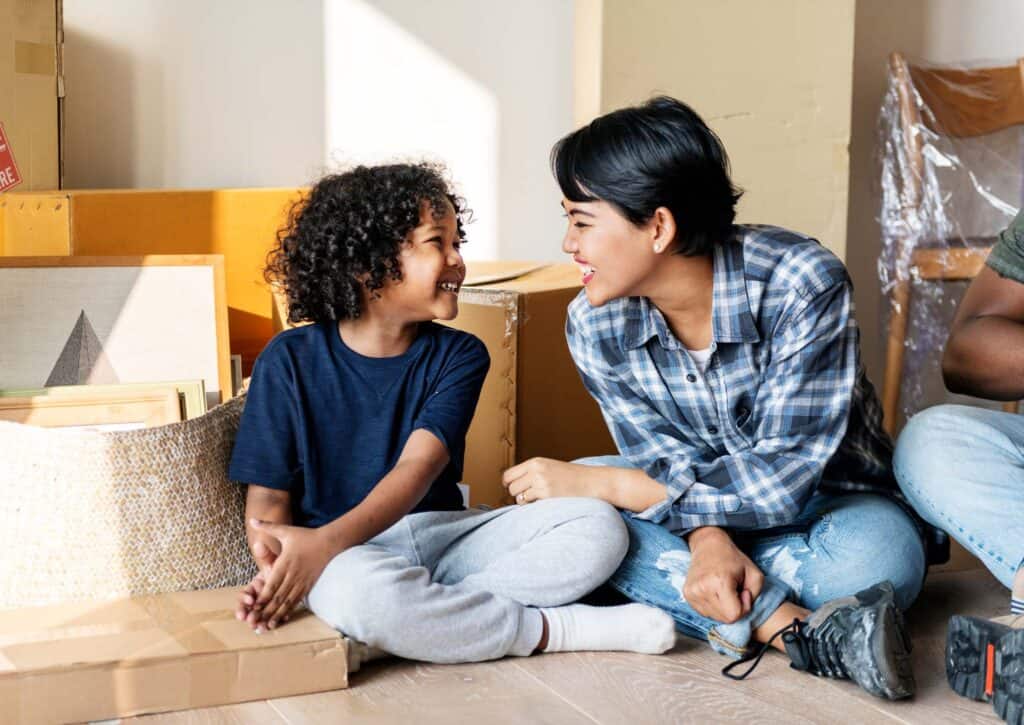 Adult and child smiling at each other while sitting beside moving boxes in a new home.