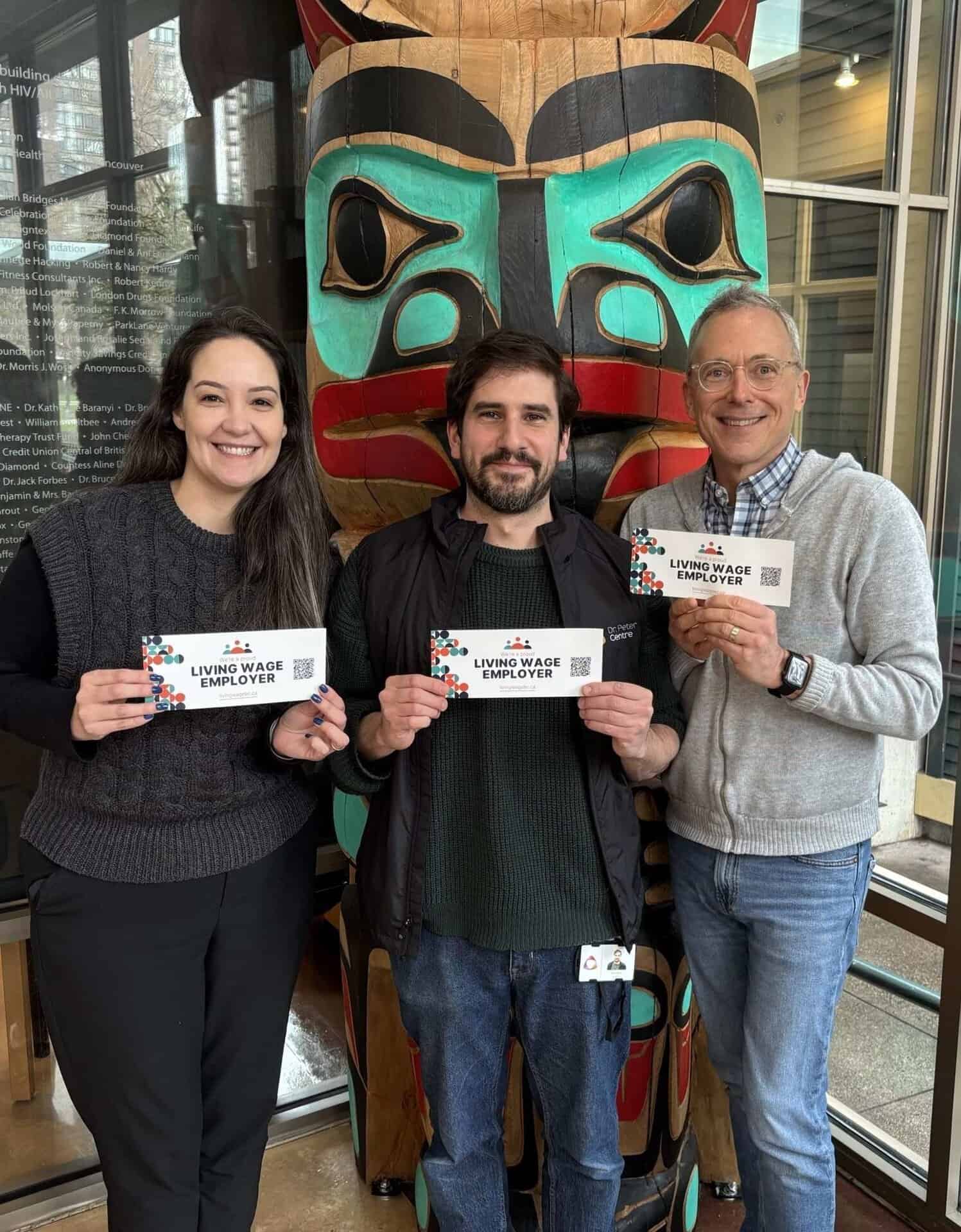 Three people holding “Living Wage Employer” cards standing in front of a large Indigenous totem pole.