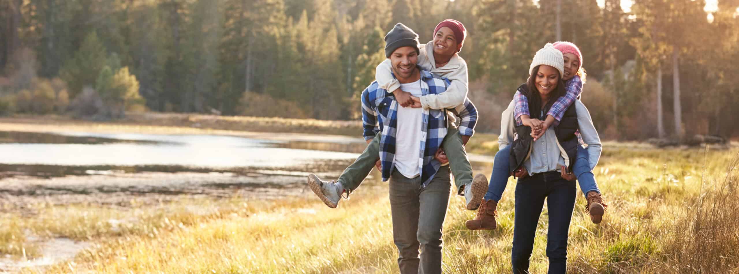 Two parents carrying their children on their backs while walking beside a lake in a forest.