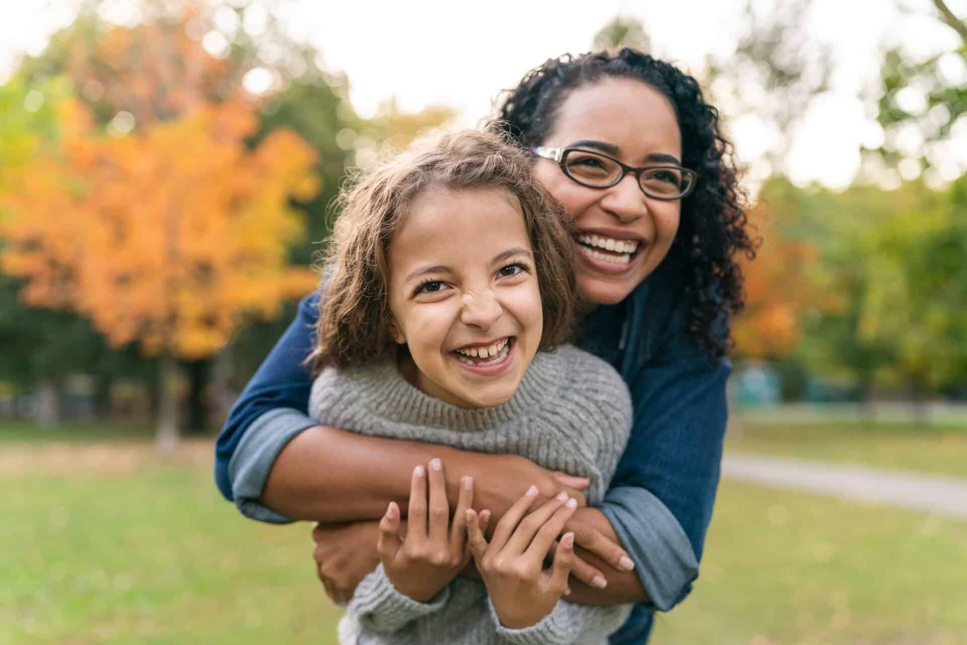Lady hugging a smiling child outdoors in a park with autumn trees.