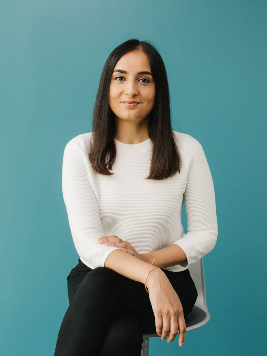Woman with long dark hair wearing a white sweater, seated on a chair against a teal background