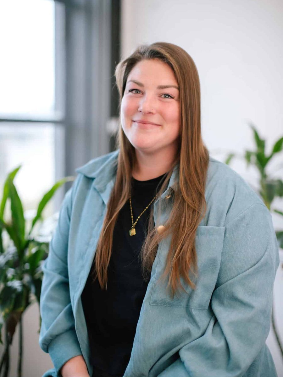 Smiling woman with long brown hair wearing a teal jacket seated in a bright office, with plants in the background