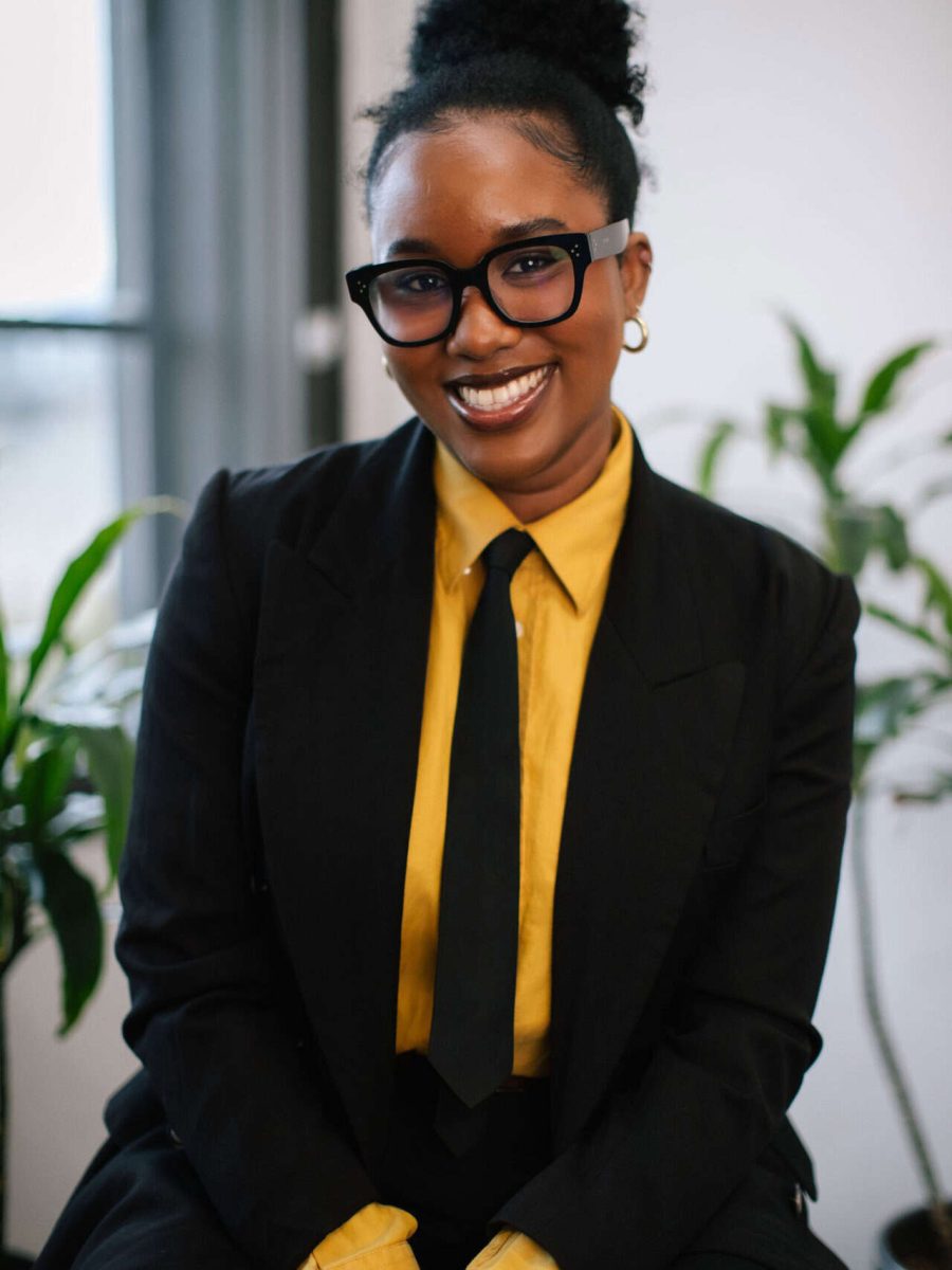 Smiling woman wearing glasses, a black blazer, and yellow shirt seated indoors in an office