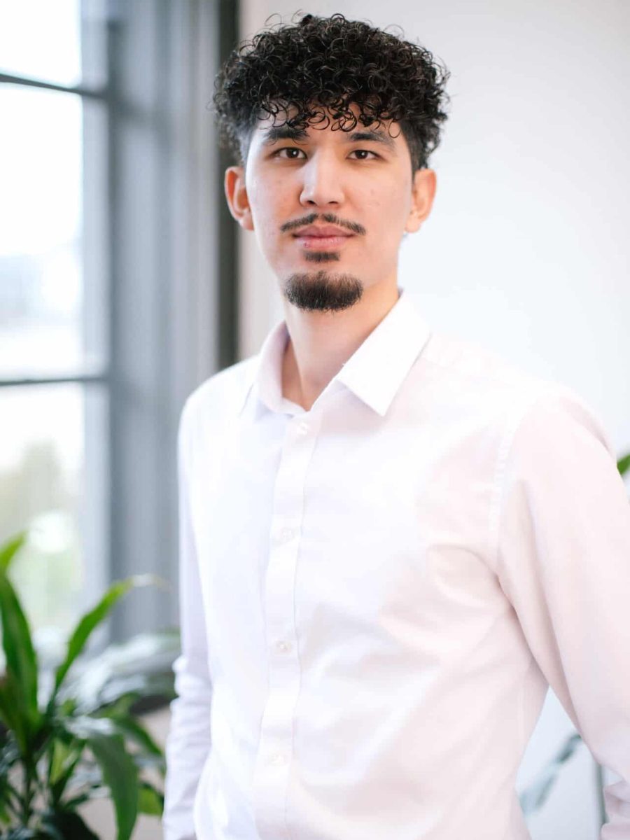 Professional headshot of a man with curly hair and a trimmed beard wearing a white shirt, standing indoors near a window