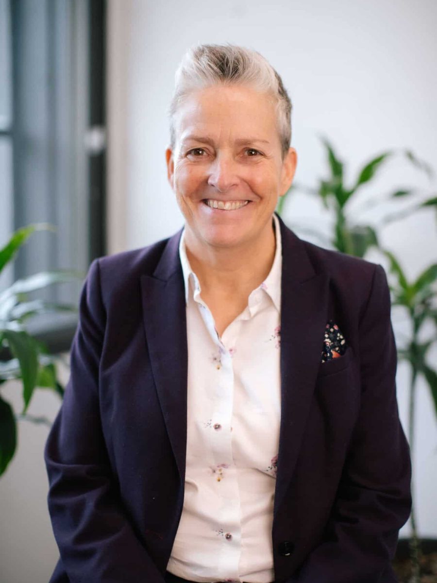 Smiling person with short gray hair wearing a purple blazer seated in a bright office with plants in the background