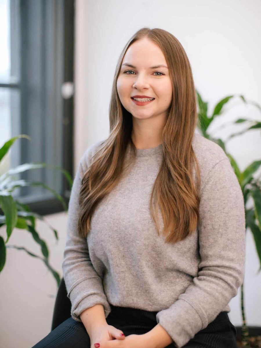 Smiling woman with long light brown hair wearing a gray sweater seated in a bright office