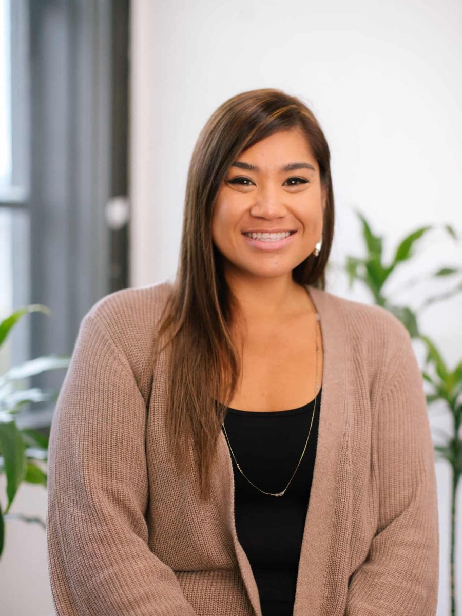 Smiling woman wearing a tan cardigan over a black top seated in a bright office with plants in the background