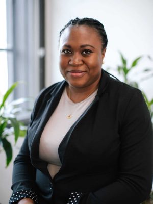 Smiling woman with braided hair wearing a black blazer seated in a bright office, with plants in the background