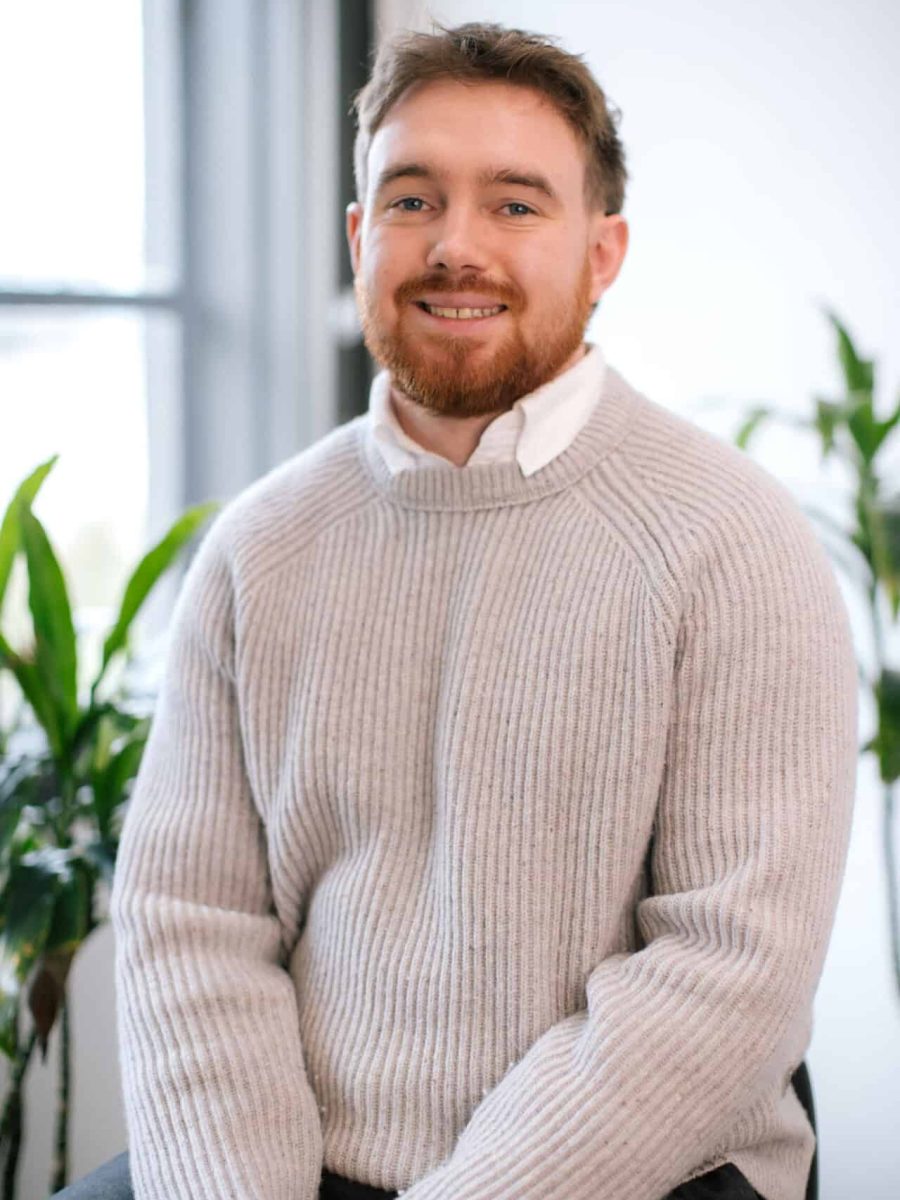 Smiling man with short hair and beard wearing a light knit sweater seated in a bright office, with plants in the background