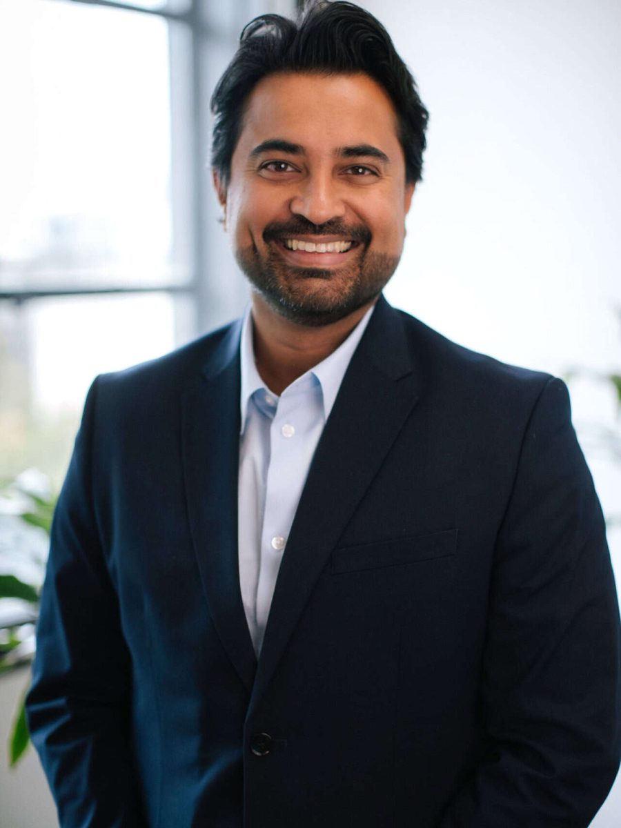 Smiling man wearing a navy blazer and light blue shirt standing in a bright office with plants in the background