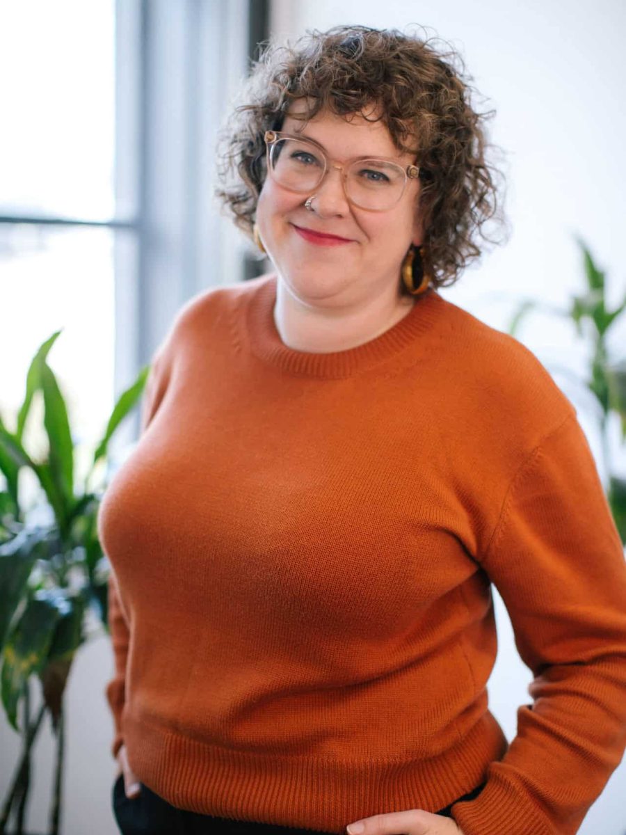Smiling woman with curly hair and glasses wearing an orange sweater in a bright office