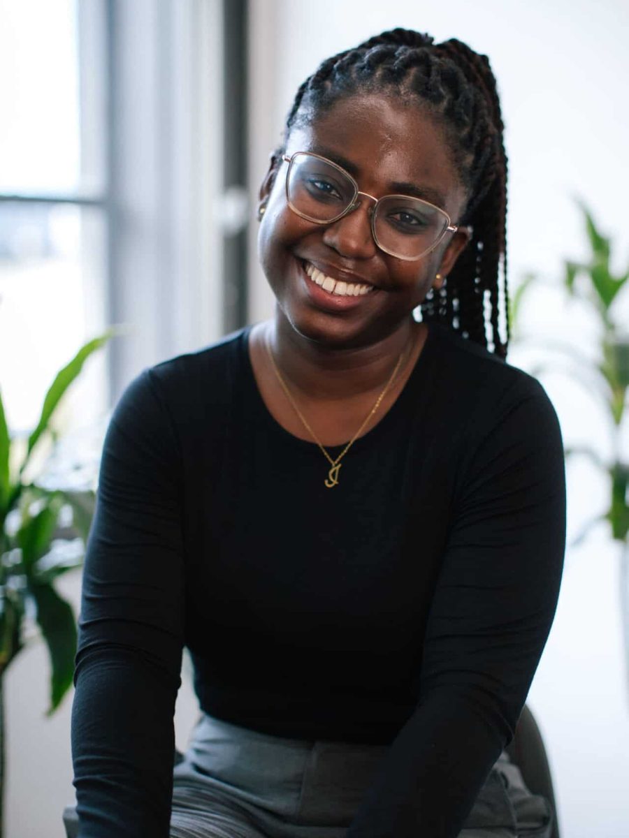 Smiling woman with braided hair and glasses wearing a black top seated in a bright office, with plants in the background
