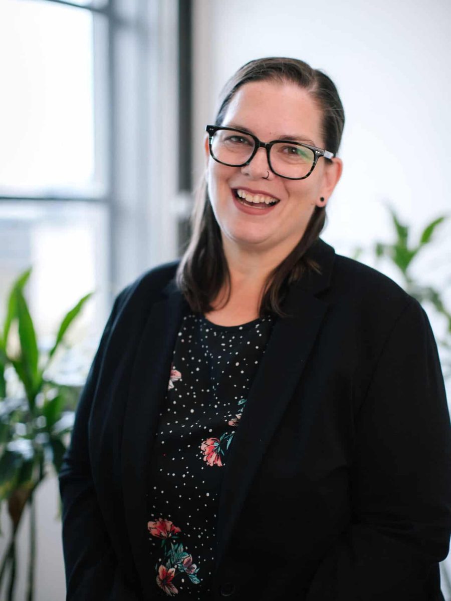 Smiling woman wearing glasses and a black blazer standing in a bright office with plants in the background