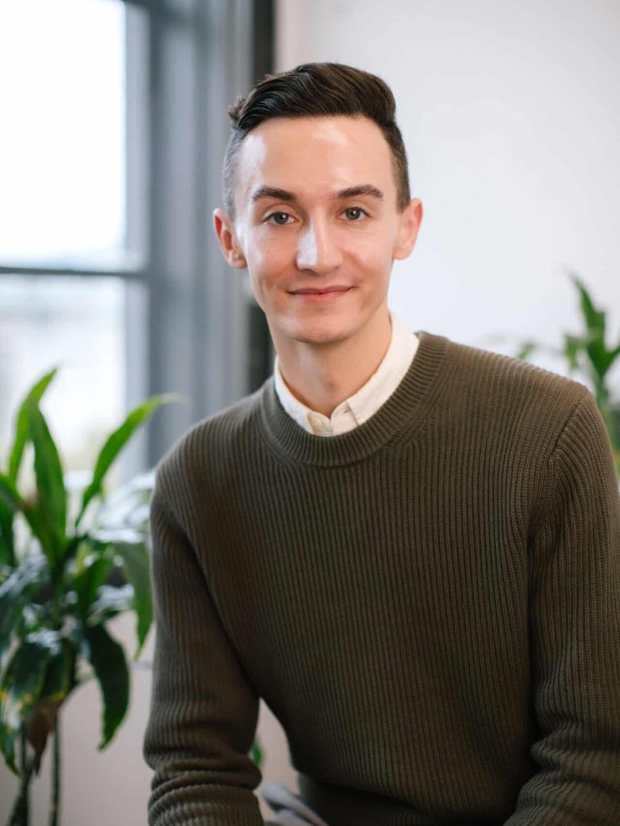 Young man wearing a green knit sweater seated in a bright office, with plants in the background