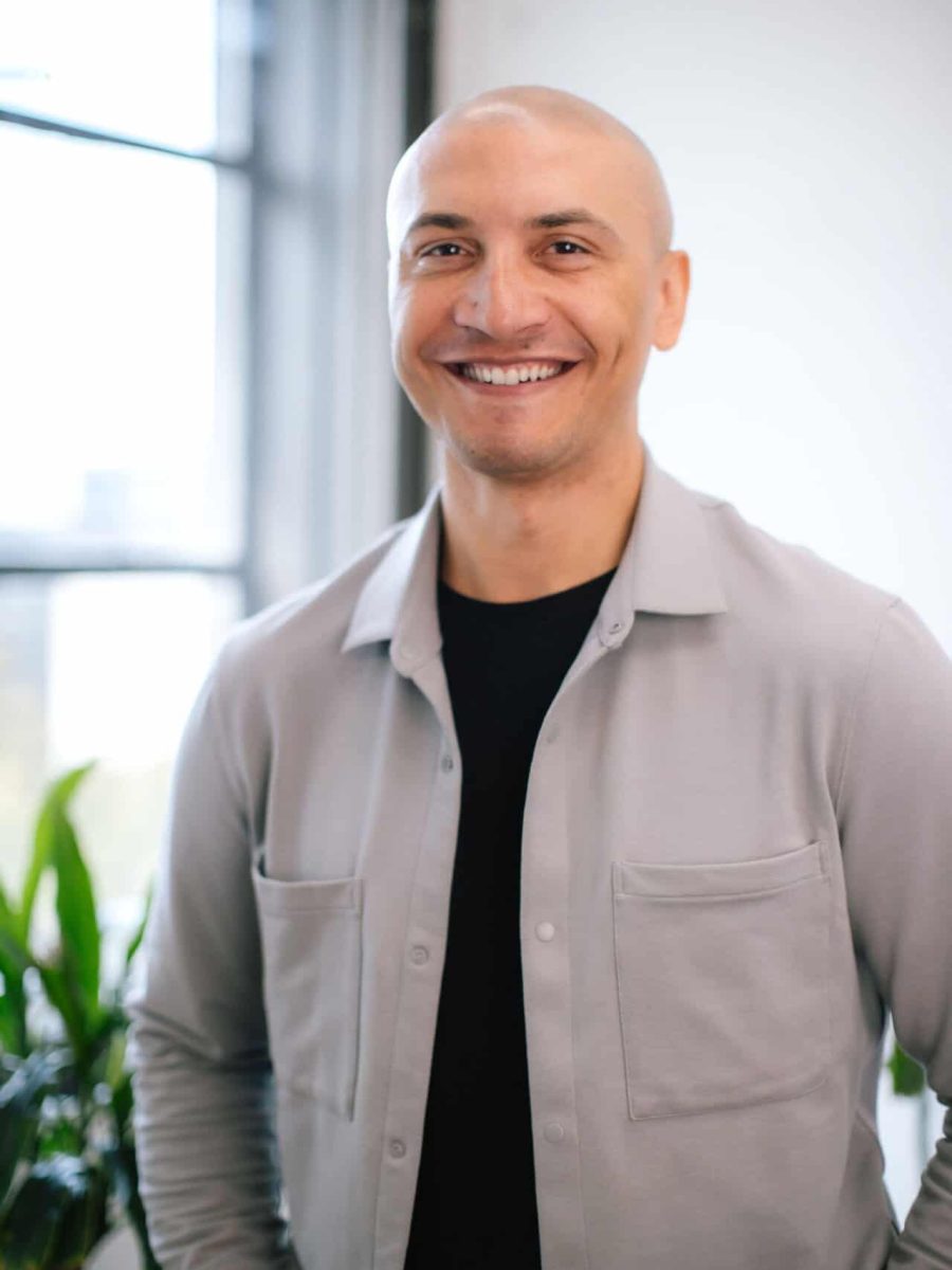 Smiling bald man wearing a light gray shirt standing in a bright office with plants in the background