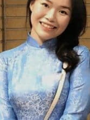 Smiling woman in a light blue traditional dress standing in front of a brick wall.