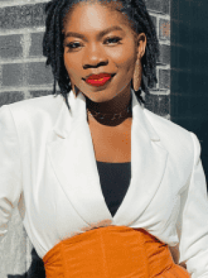 Woman with braided hair wearing a white blazer standing by a brick wall.