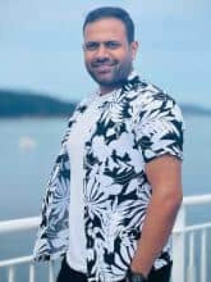 Man smiling by a waterfront railing wearing a black-and-white floral shirt.
